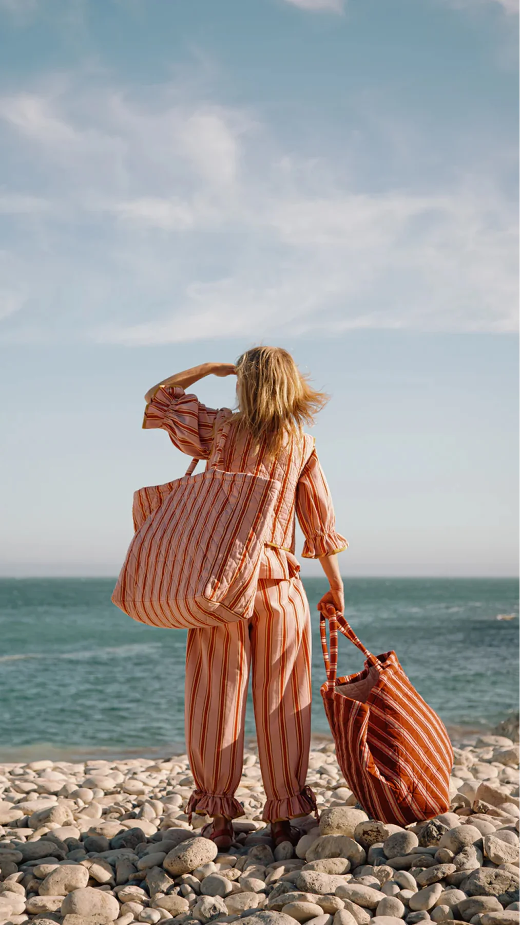 Person wearing a pink striped outfit stands on a pebble beach facing the ocean, carrying a matching striped tote bag over the shoulder.