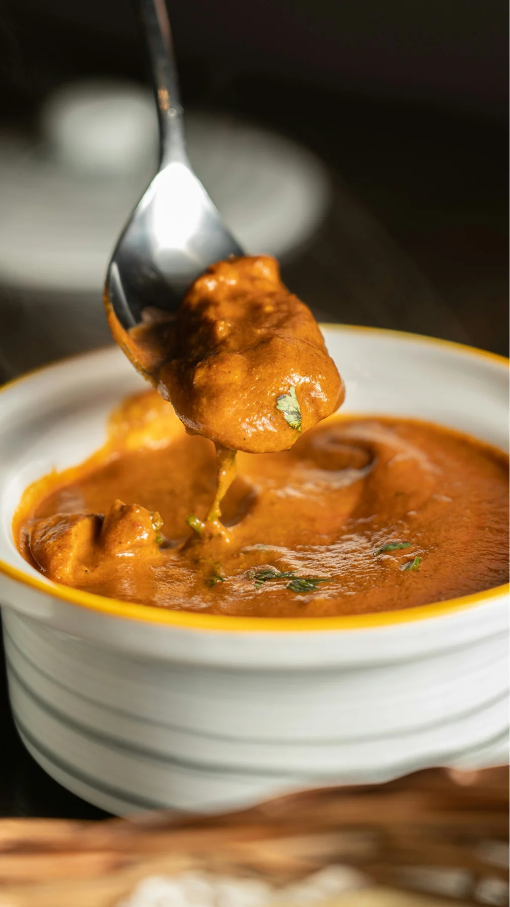 Spoon lifting a piece of chicken from a creamy orange curry in a white bowl.