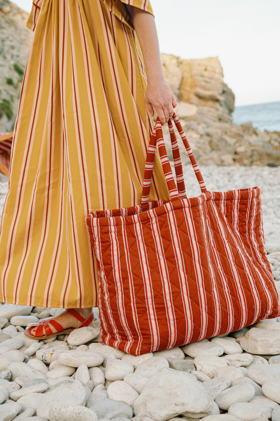 Person in a mustard yellow striped dress carries a large red striped tote bag on a rocky beach.