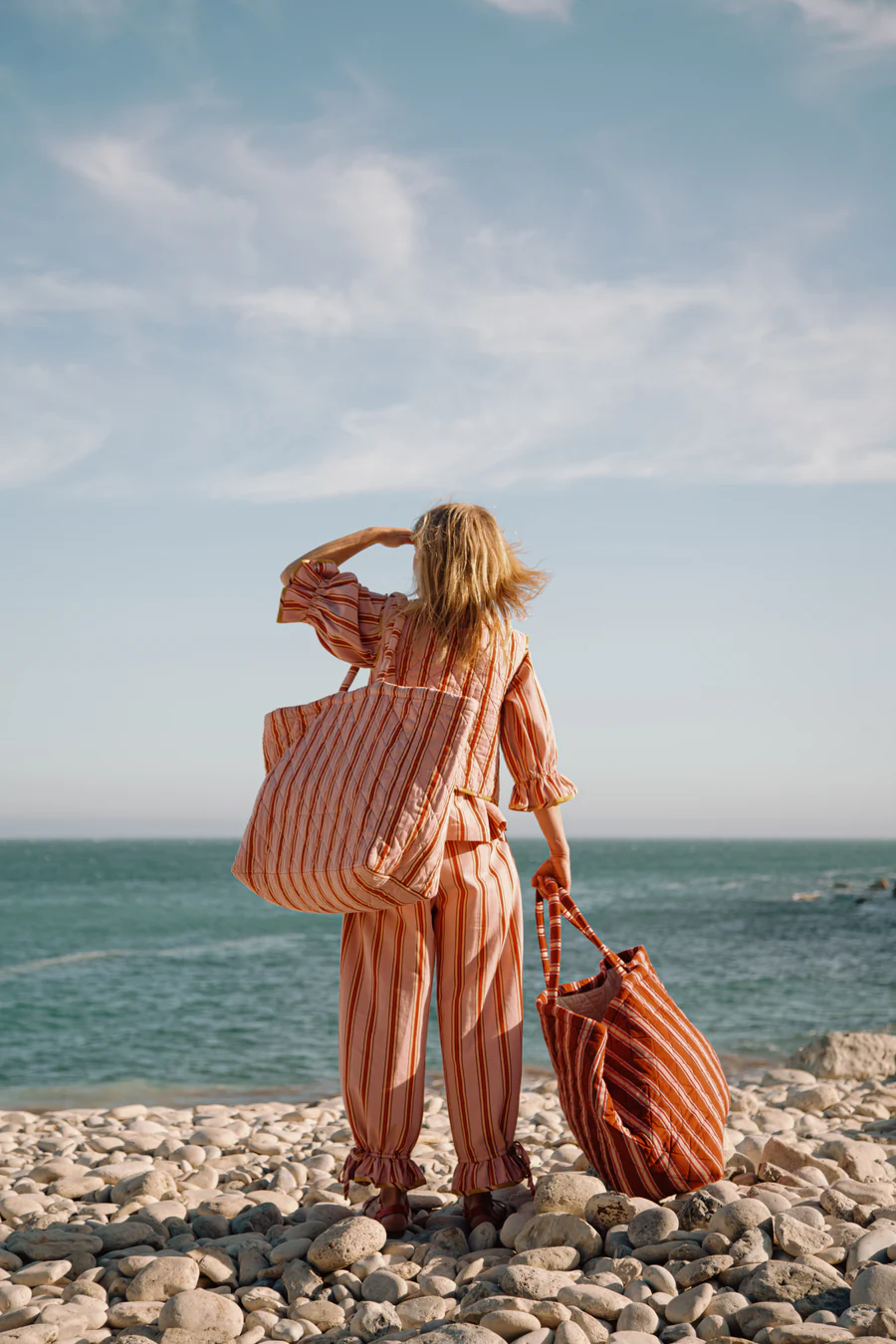 Back view of a person standing on a pebbled beach, wearing a peach-pink striped outfit and carrying a matching striped tote bag while facing the ocean.