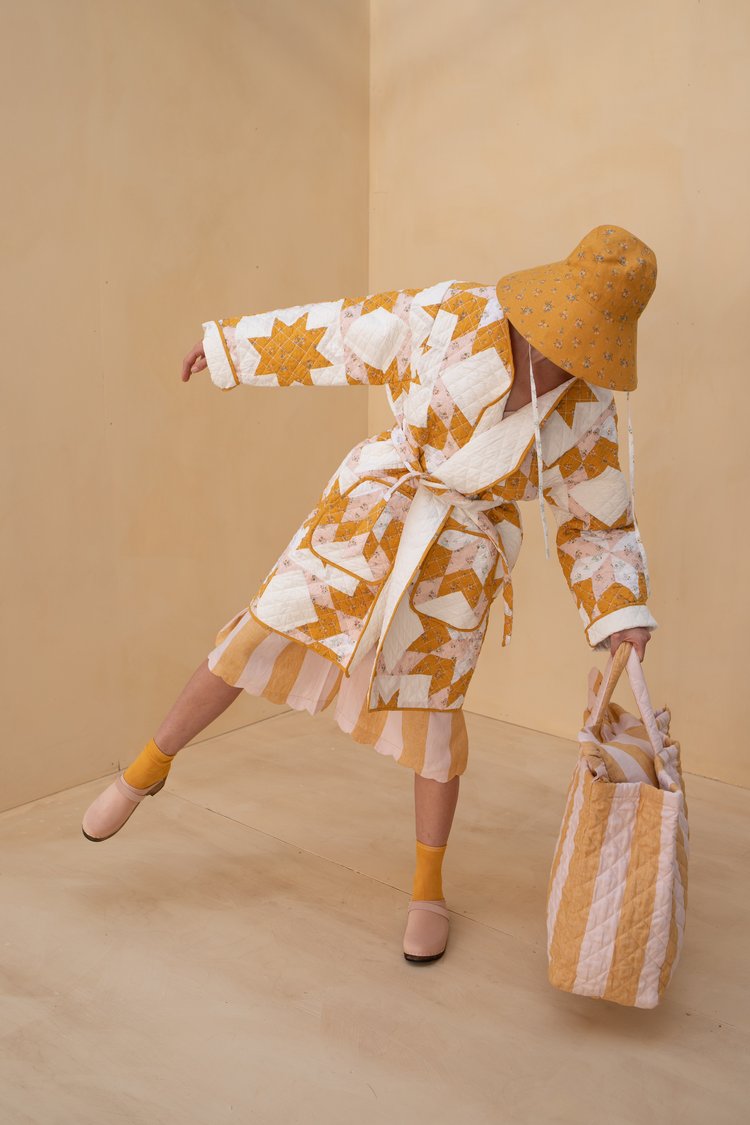 Person posing in a yellow-and-white quilted coat with a matching skirt and wide-brimmed yellow hat, holding a striped tote bag