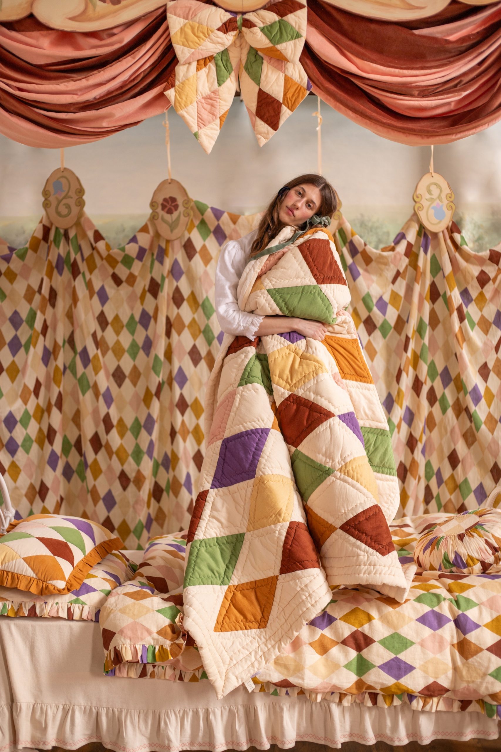 Young woman wrapped in a multicolor patchwork quilt on a stage with pink draped curtains behind her.