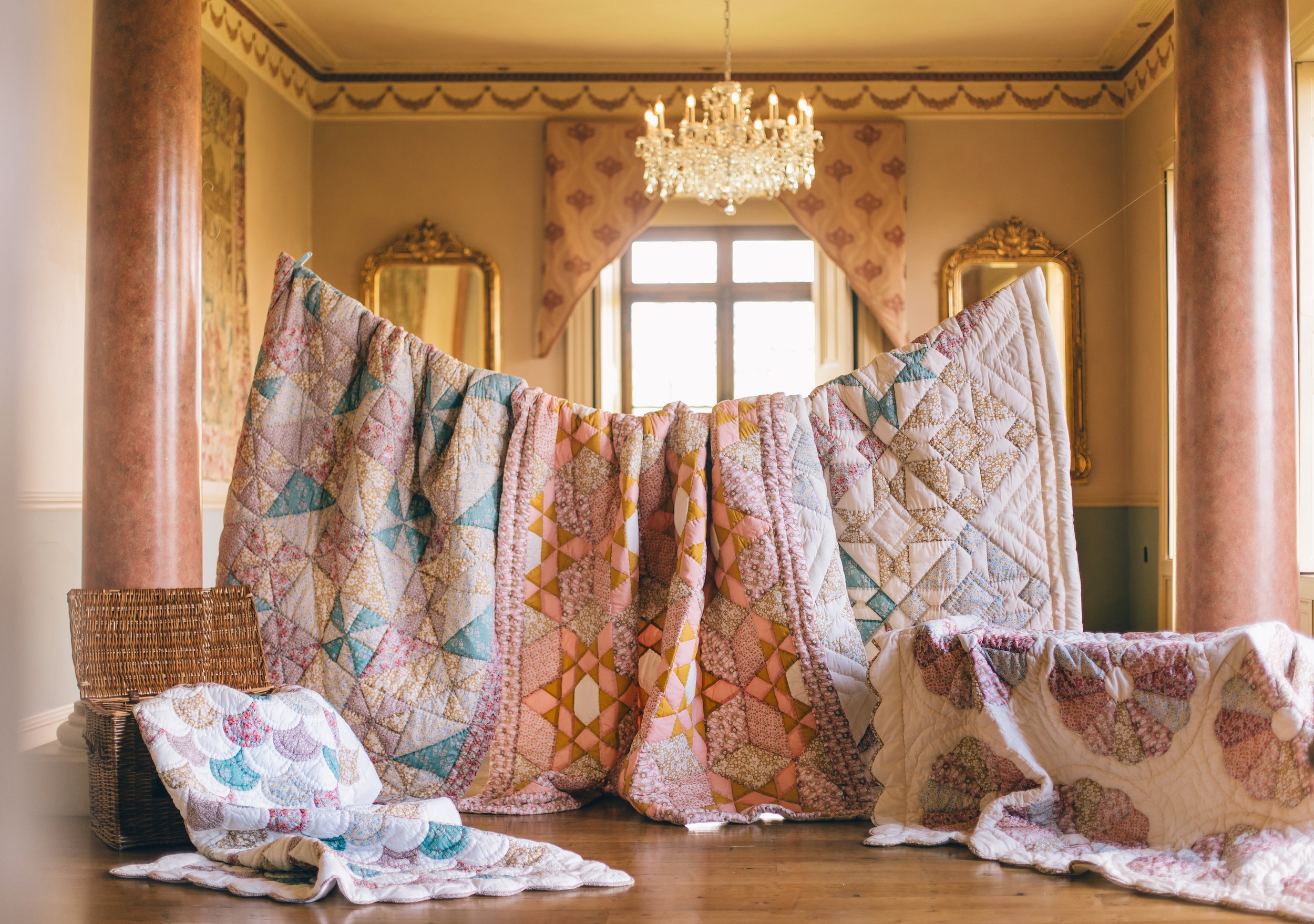 Quilted pastel patchwork blankets are draped like a screen across a room, with a crystal chandelier and ornate mirrors in the background.
