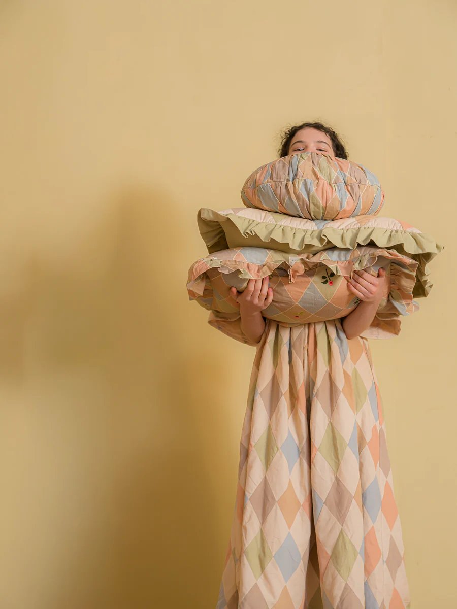 Person in a pastel argyle dress hugs a large stack of pillows in front of a yellow wall, peeking over them.