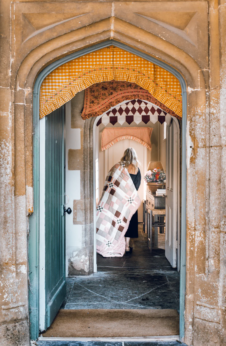 A woman wrapped in a patchwork quilt walks through a stone arched doorway into a cozy, fabric-filled room.